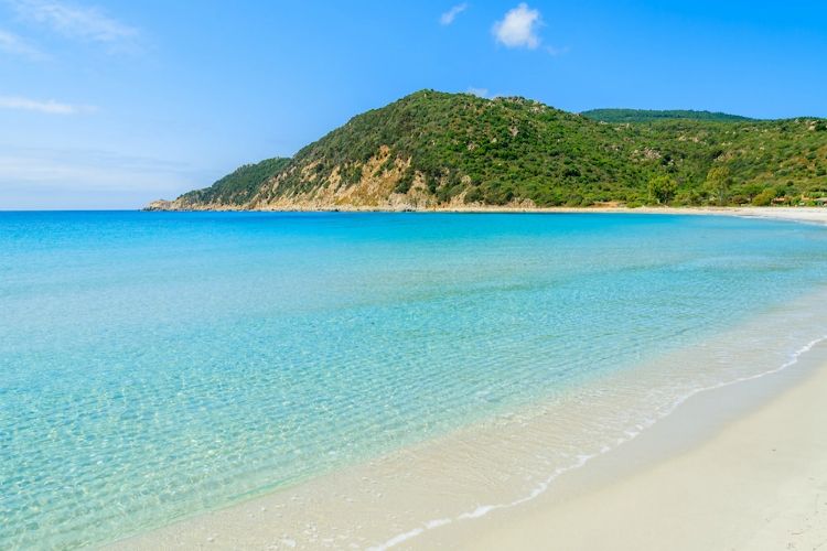 beaches in sardinia | blue water and sand beach under clear skies with green hillside in distance | Cala Pira