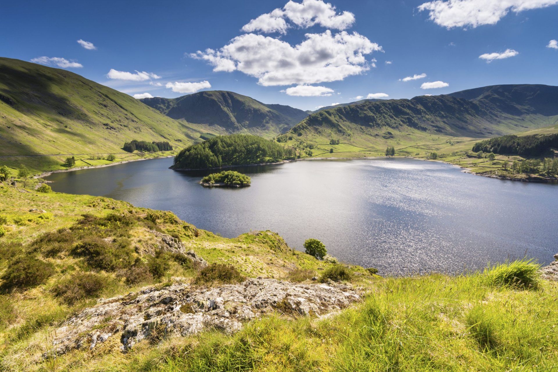 Haweswater Reservoir in Mardale Valley taken from Whiteacre Crag. A green hill that leads down to the water is in the foreground. The water is deep blue and reflects the sunlight. The reservoir is surrounded by rolling green hills, and a deep blue sky filled with fluffy white clouds is overhead