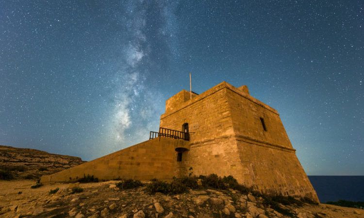 A beautiful Milky Way night time photograph of the Dwejra Watch Tower on the island of Gozo, Malta. With the orange building in the foreground and the stars against a deep blue sky.