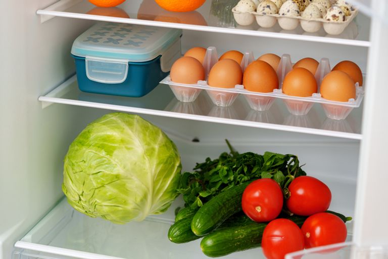 A white refrigerator is open, revealing a well-stocked interior with fresh fruits, vegetables, eggs, and other groceries.